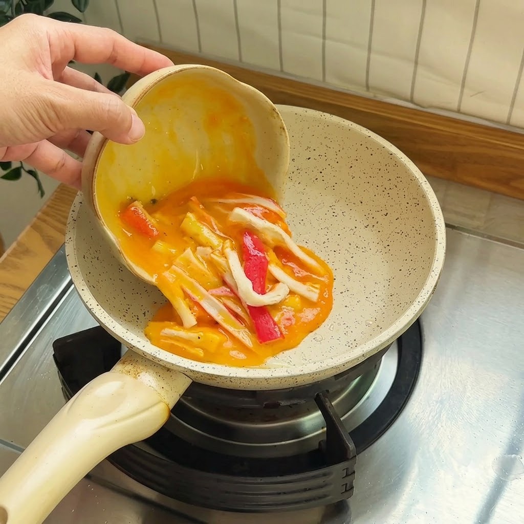 Roztrzepane jajka z kawałkami czerwono-białych paluszków surimi wylewane na małą patelnię z powłoką nieprzywierającą.