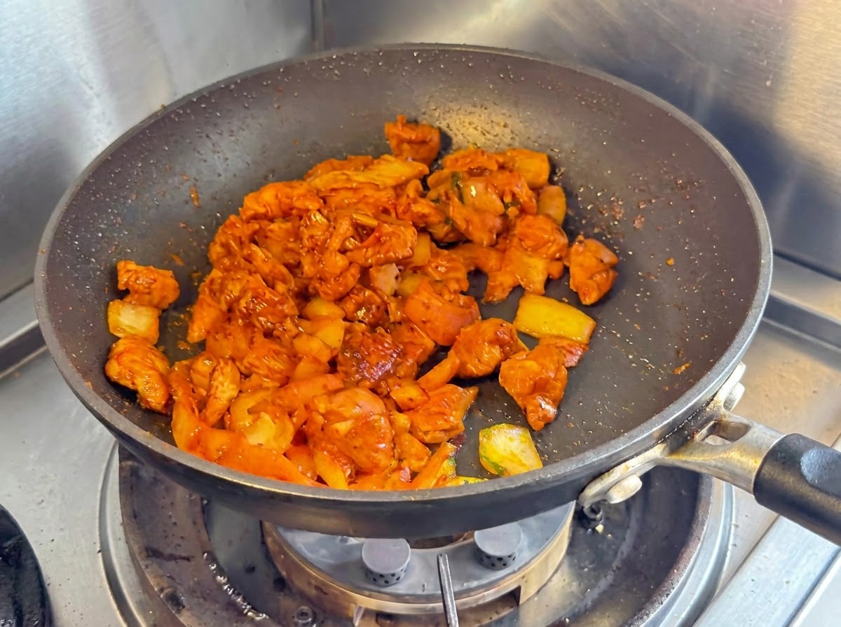 Pokrojoną pierś kurczaka, cebulę i posiekane kimchi smażone razem na ciemnej patelni nieprzywierającej.