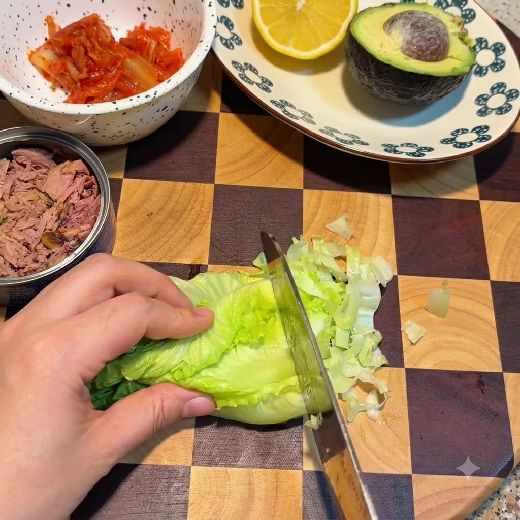 Nóż krojący świeże liście zielonej sałaty na desce obok miseczki z kimchi i puszki tuńczyka.