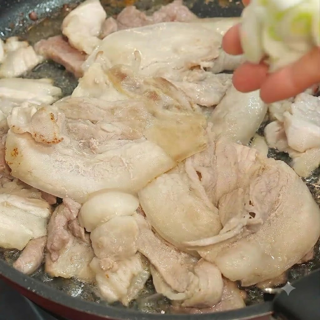 Ręka wrzucająca posiekaną zieloną cebulkę do patelni wypełnionej smażącymi się plastrami wieprzowiny.