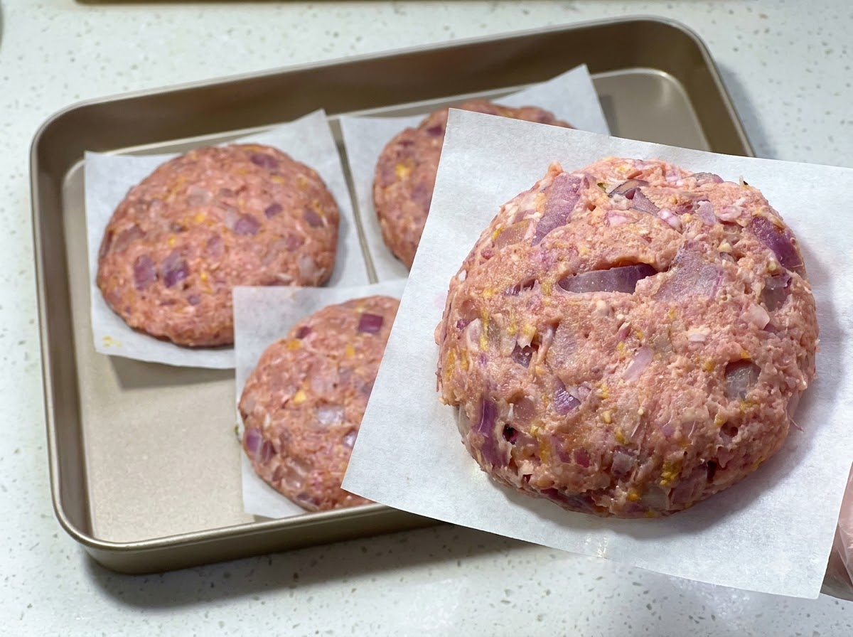 Świeżo uformowany surowy kotlet mielony na papierze do pieczenia, z innymi kotletami ułożonymi na tacy w tle.