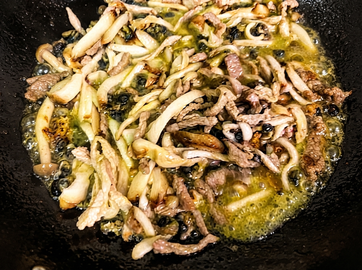 Drobno posiekane grzyby shiitake, enoki i ślimaki morskie smażące się w gorącym oleju w czarnym woku.