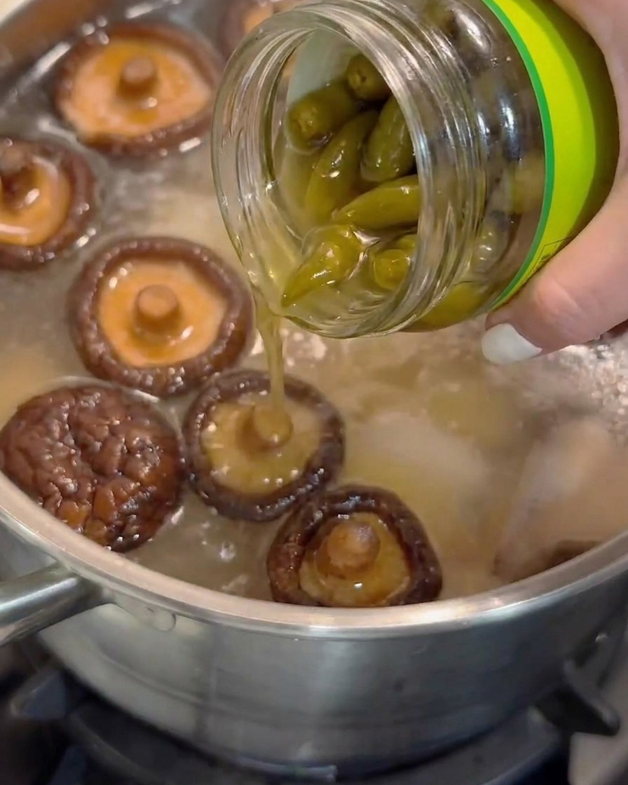 Sok z marynowanych obranych papryczek chilli wlewany ze szklanego słoika do garnka z gotującą się zupą z grzybami shiitake.