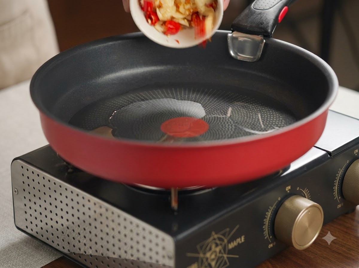 Rozgnieciony czosnek i czerwone chili wsypywane z małego białego naczynia na rozgrzaną czerwoną patelnię z olejem.