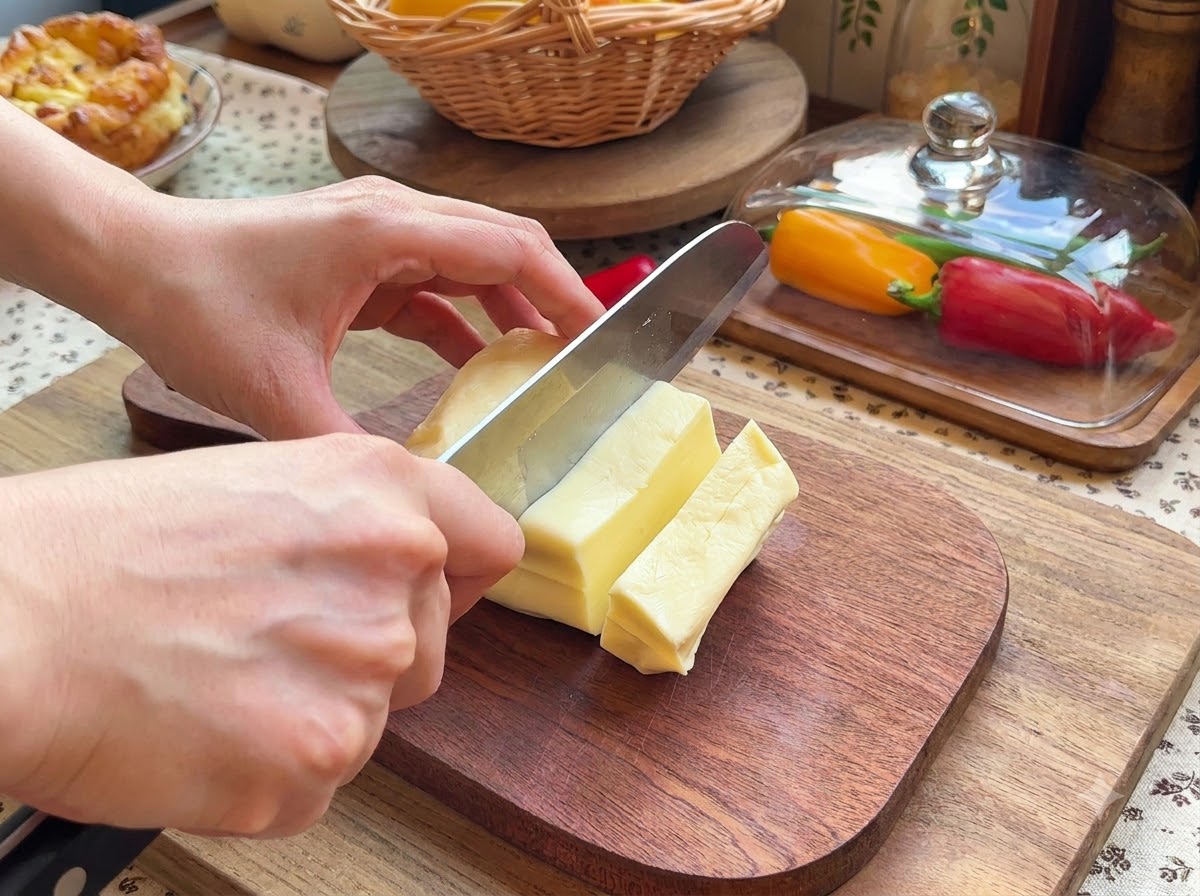 Dwie dłonie używające noża do krojenia bloku sera mozzarella na małe prostokątne kawałki na drewnianej desce do krojenia.