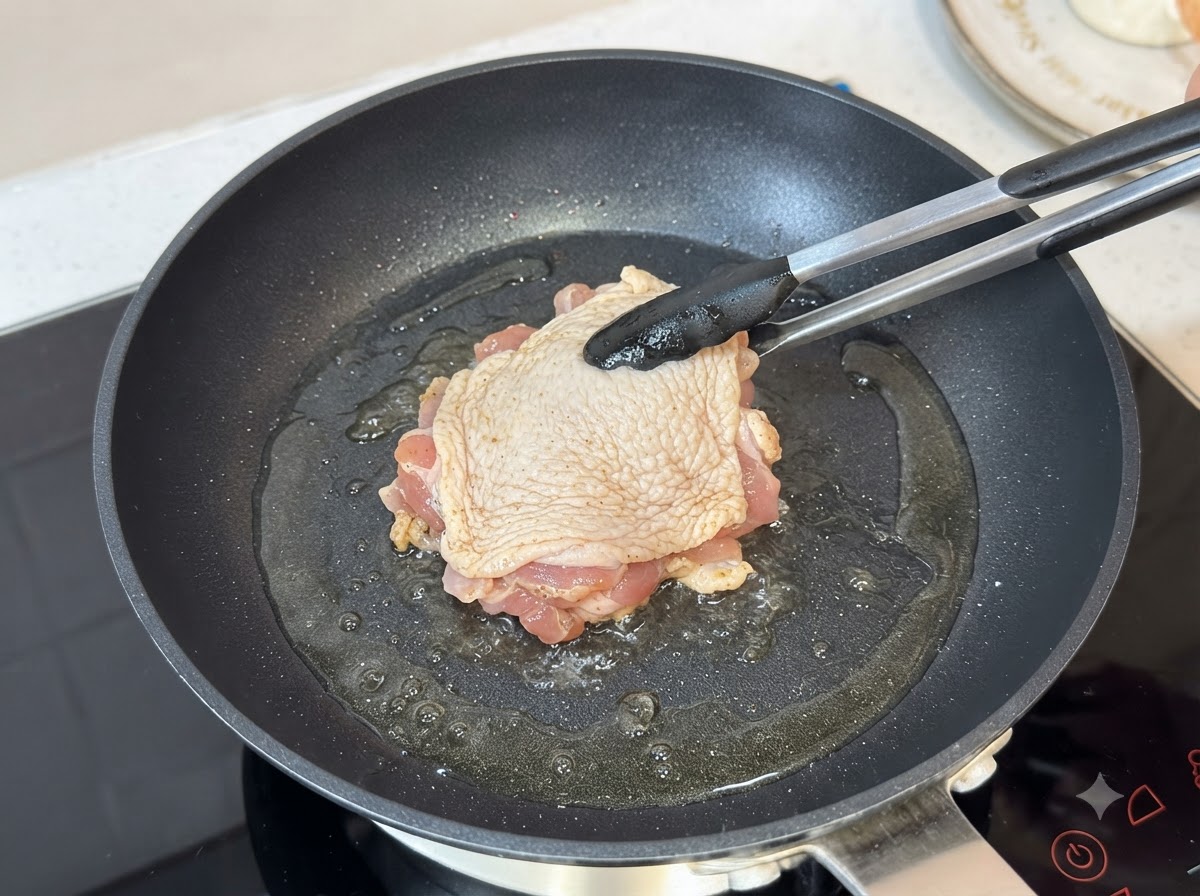 Zamarynowane udko z kurczaka smażone skórą do dołu na rozgrzanej patelni za pomocą szczypiec kuchennych.