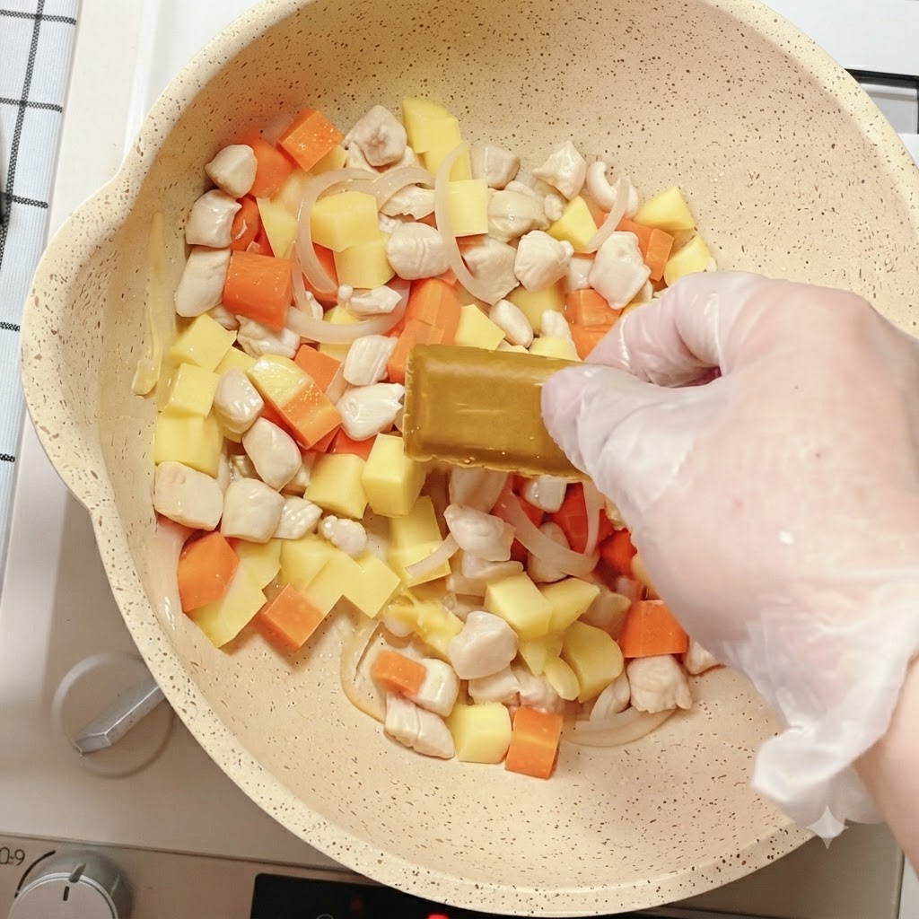 Dłoń umieszczająca kostkę przyprawy curry w patelni wypełnionej mieszanką warzyw i kurczaka.