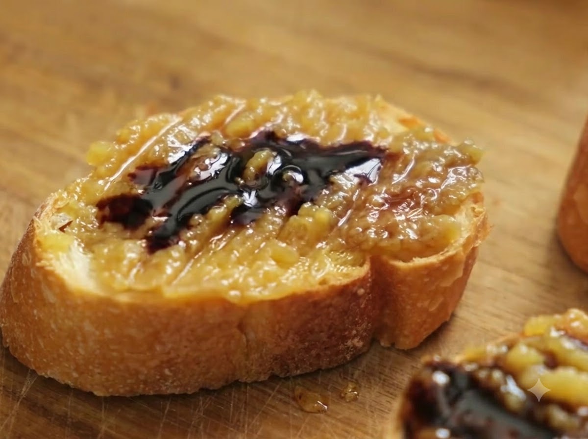Zbliżenie na kromkę bagietki posmarowaną pastą confit z czosnku i octem balsamicznym, posypaną gruboziarnistą solą morską.