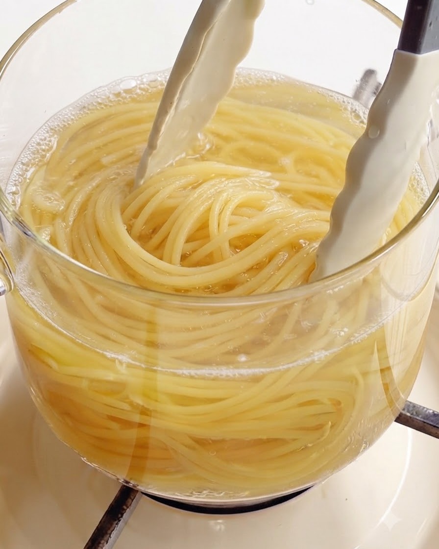 Używanie białych szczypiec kuchennych do wyjmowania ugotowanego spaghetti ze szklanego garnka z wrzącą wodą.