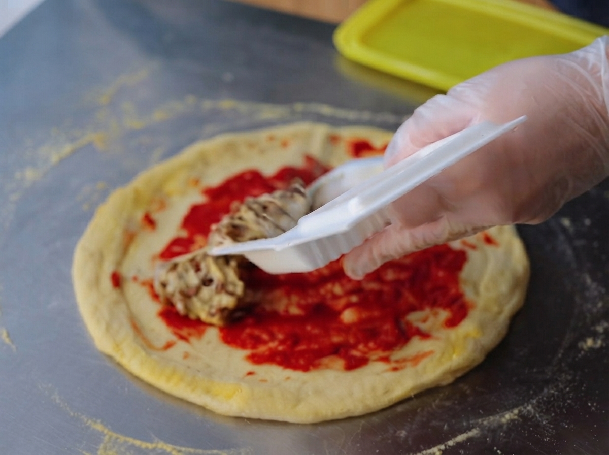 Ręka w rękawiczce rozprowadzająca lepkie, ciągnące się natto równomiernie na warstwie jasnoczerwonej pasty pomidorowej na surowym cieście do pizzy.