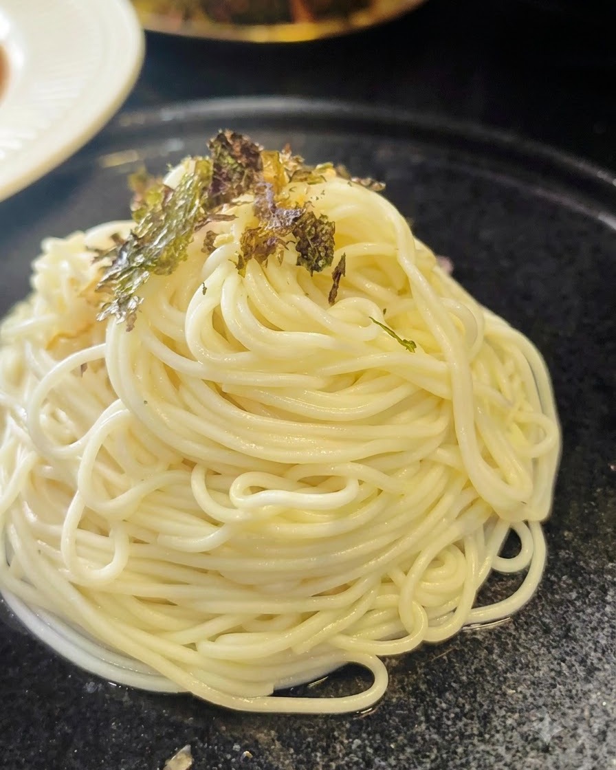 Pokruszone płatki suszonych wodorostów posypywane na schludnie ułożoną spiralę ugotowanego makaronu ramen na ciemnym talerzu.
