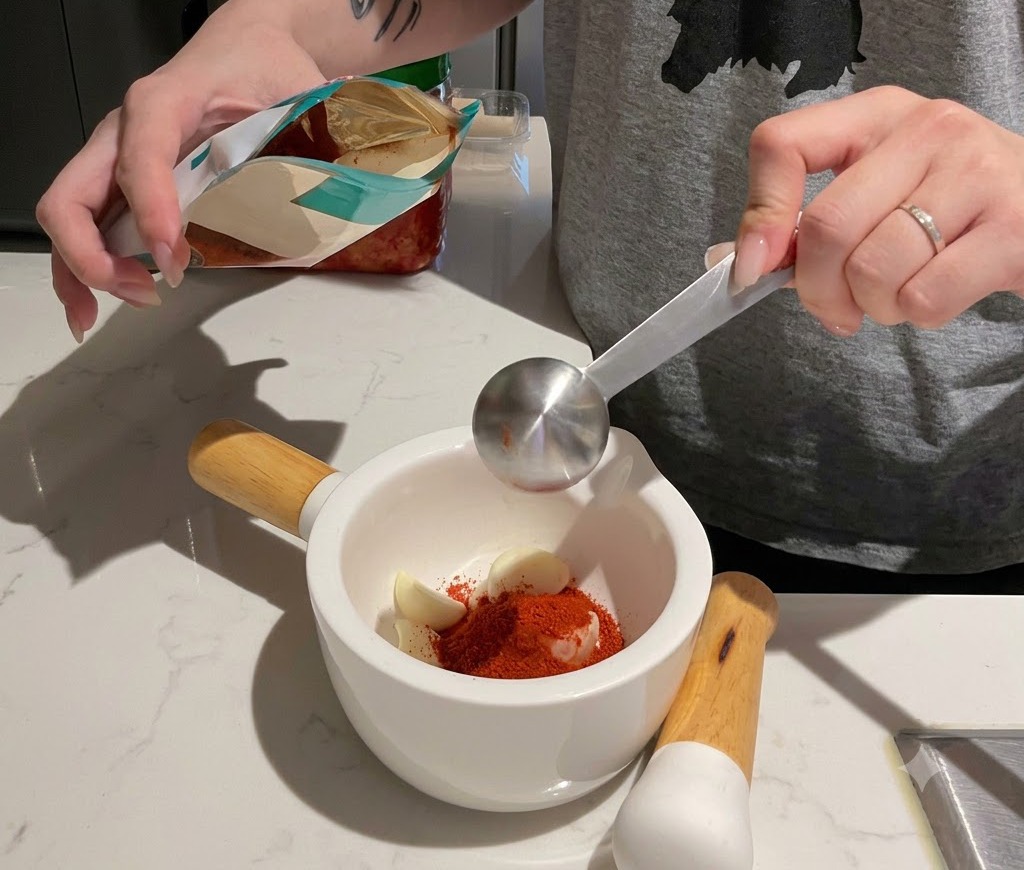 Odmierzanie sproszkowanego czerwonego chili do białego moździerza wypełnionego ząbkami czosnku.