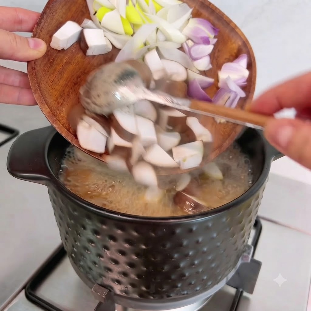 Pokrojone w kostkę grzyby shiitake, cebula i dymka zsuwane z drewnianej deski do gotującego się garnka.