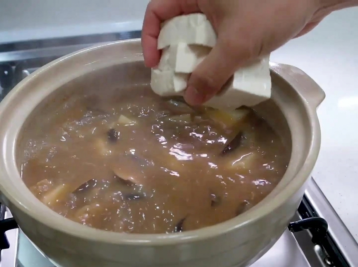Dłoń ostrożnie wrzucająca świeże, białe kostki tofu do garnka z bulgoczącym brązowym gulaszem.