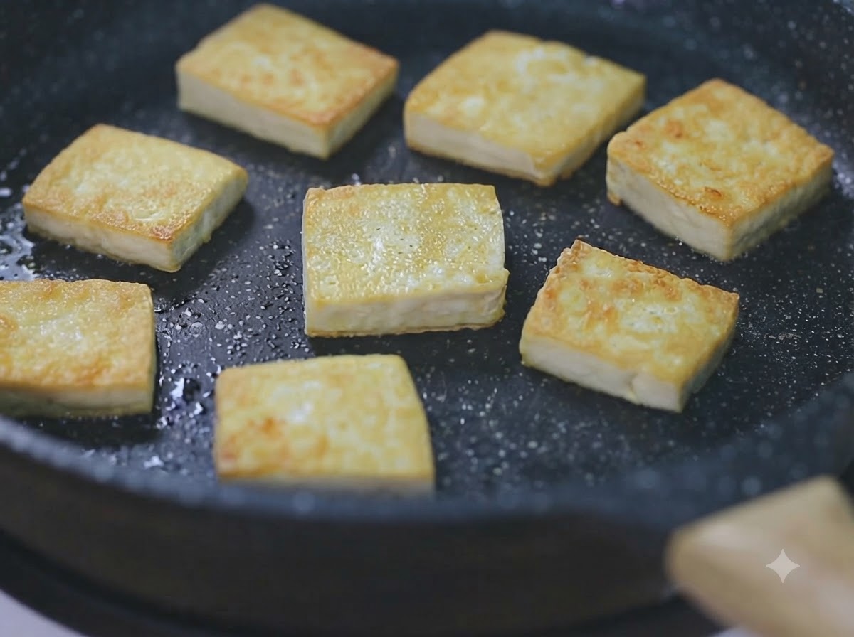 Kilka złotobrązowych kostek tofu skwierczących na czarnej patelni na małym ogniu.