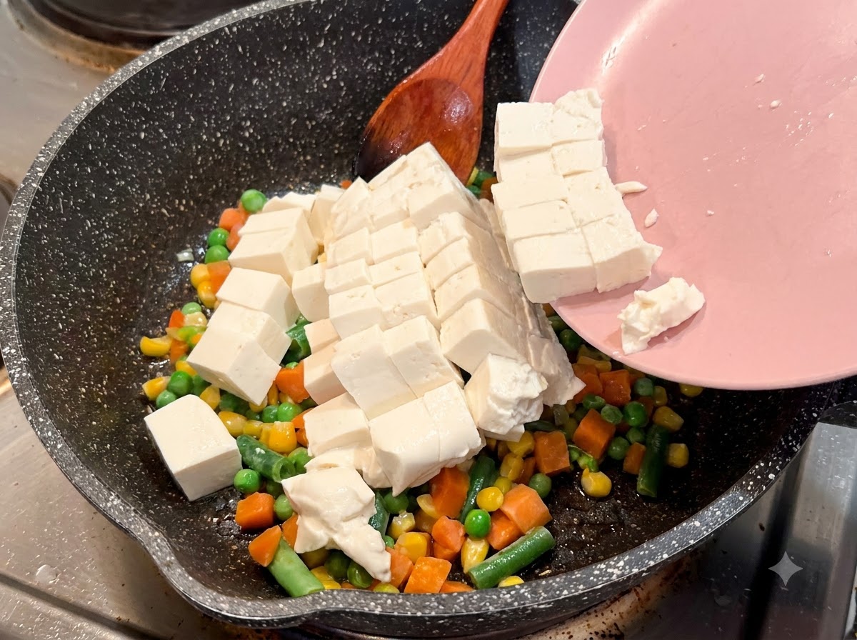 Pokrojone w kostkę miękkie tofu zsuwające się z różowego talerza na nakrapianą patelnię z warzywami.
