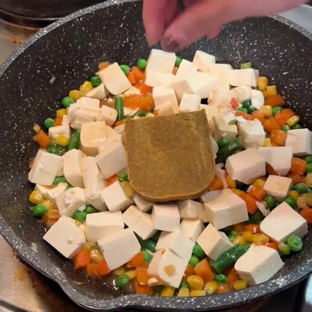 Ręka umieszczająca blok stałego brązowego roux curry na tofu i warzywach na patelni.