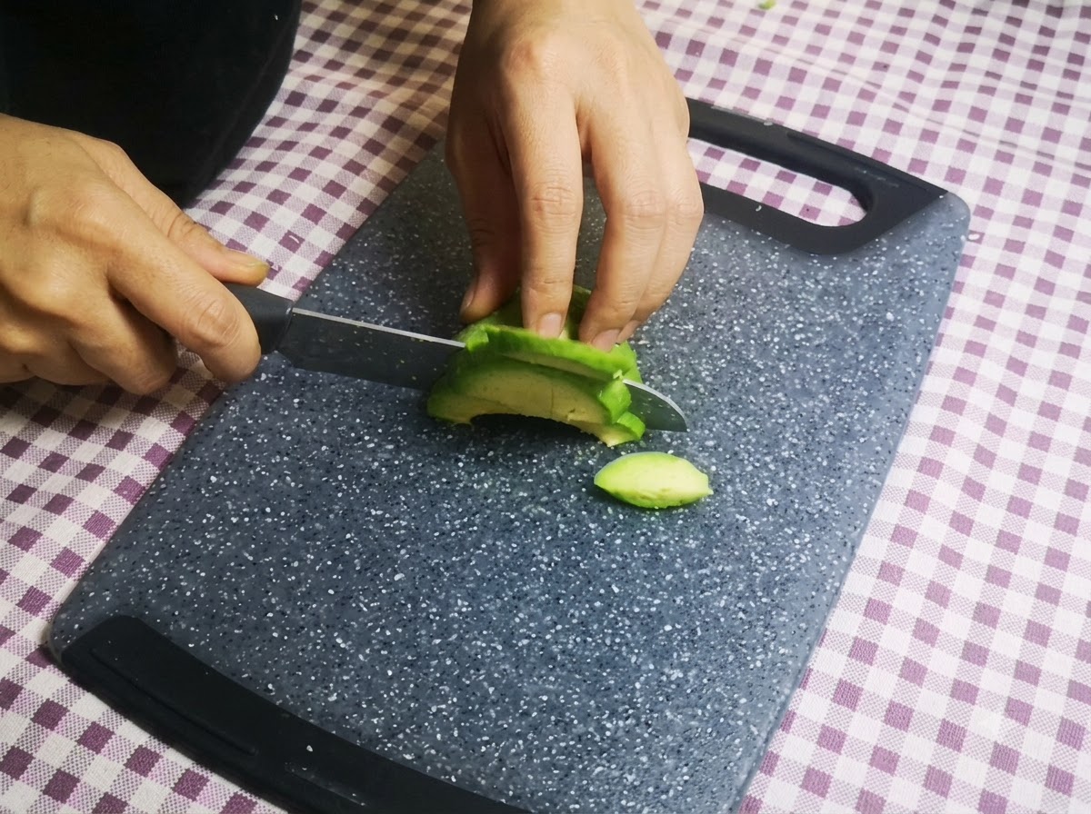 Dłonie krojące połówkę zielonego awokado na cienkie plasterki za pomocą noża na desce do krojenia.