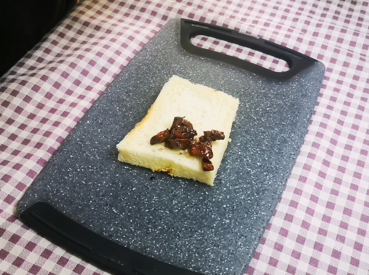 Zbliżenie na kawałki ciemnego, glazurowanego kurczaka starannie ułożone na pojedynczej kromce białego chleba leżącej na szarej desce do krojenia.