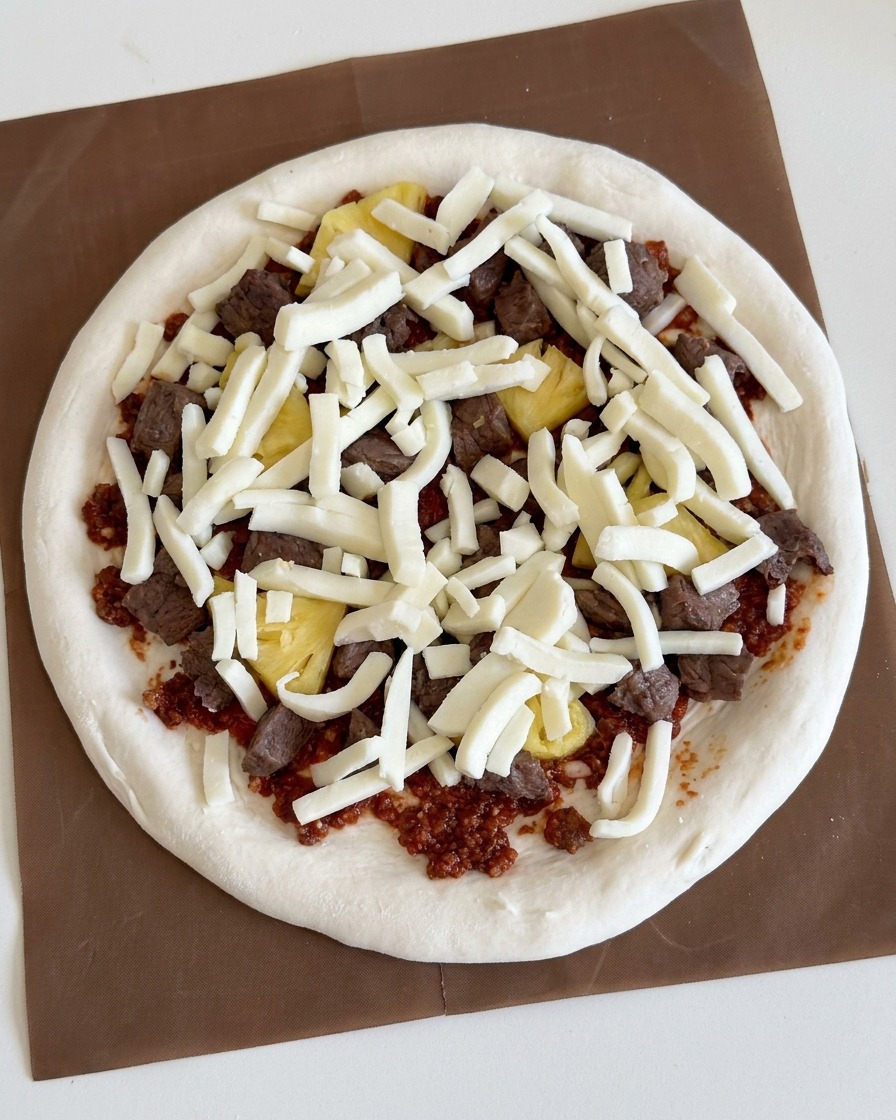 Starta, biała mozzarella obficie posypana na mięso i ananasa ułożone na pizzy.