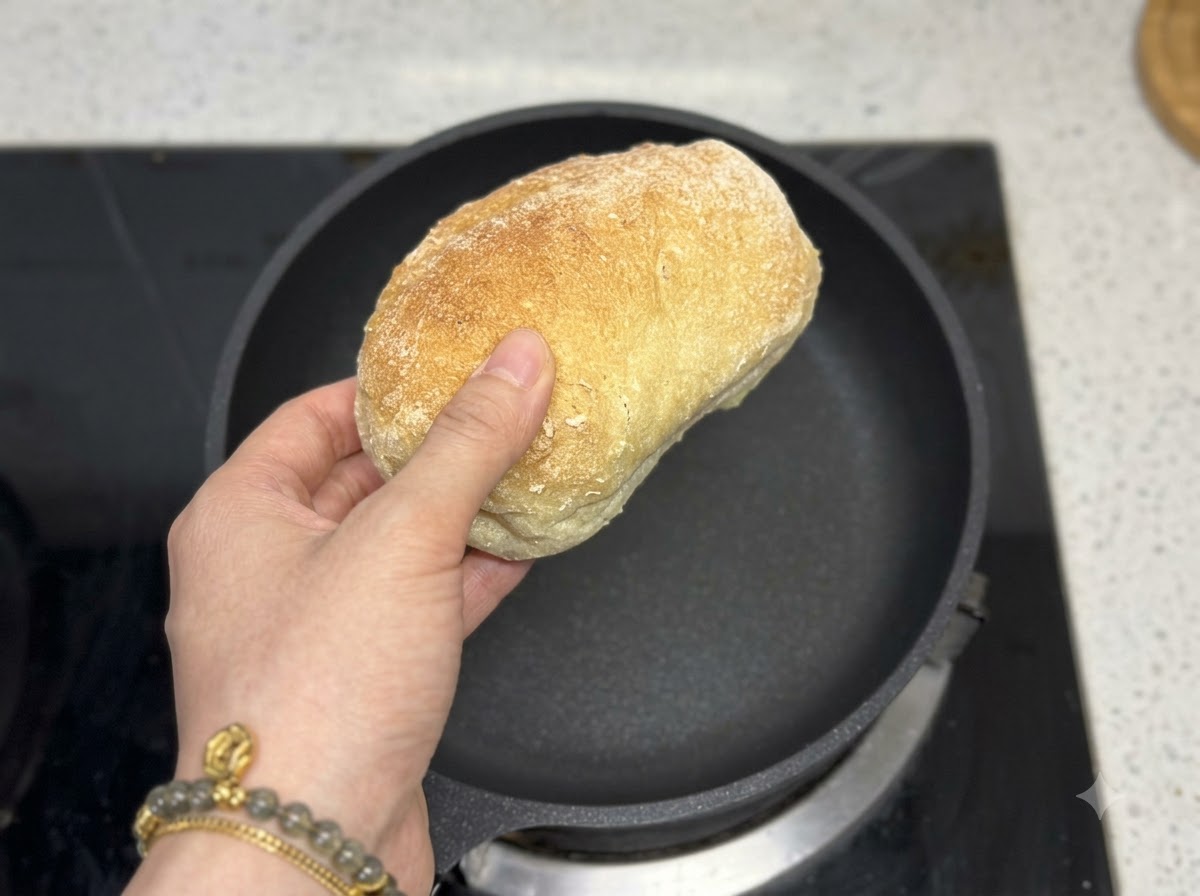 Ręka trzymająca bochenek ciabatty bezpośrednio nad pustą czarną patelnią.