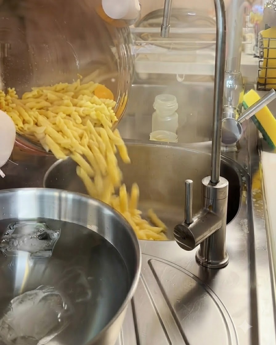 Ugotowany makaron fusilli odsączany w metalowym durszlaku nad miską z wodą z lodem.
