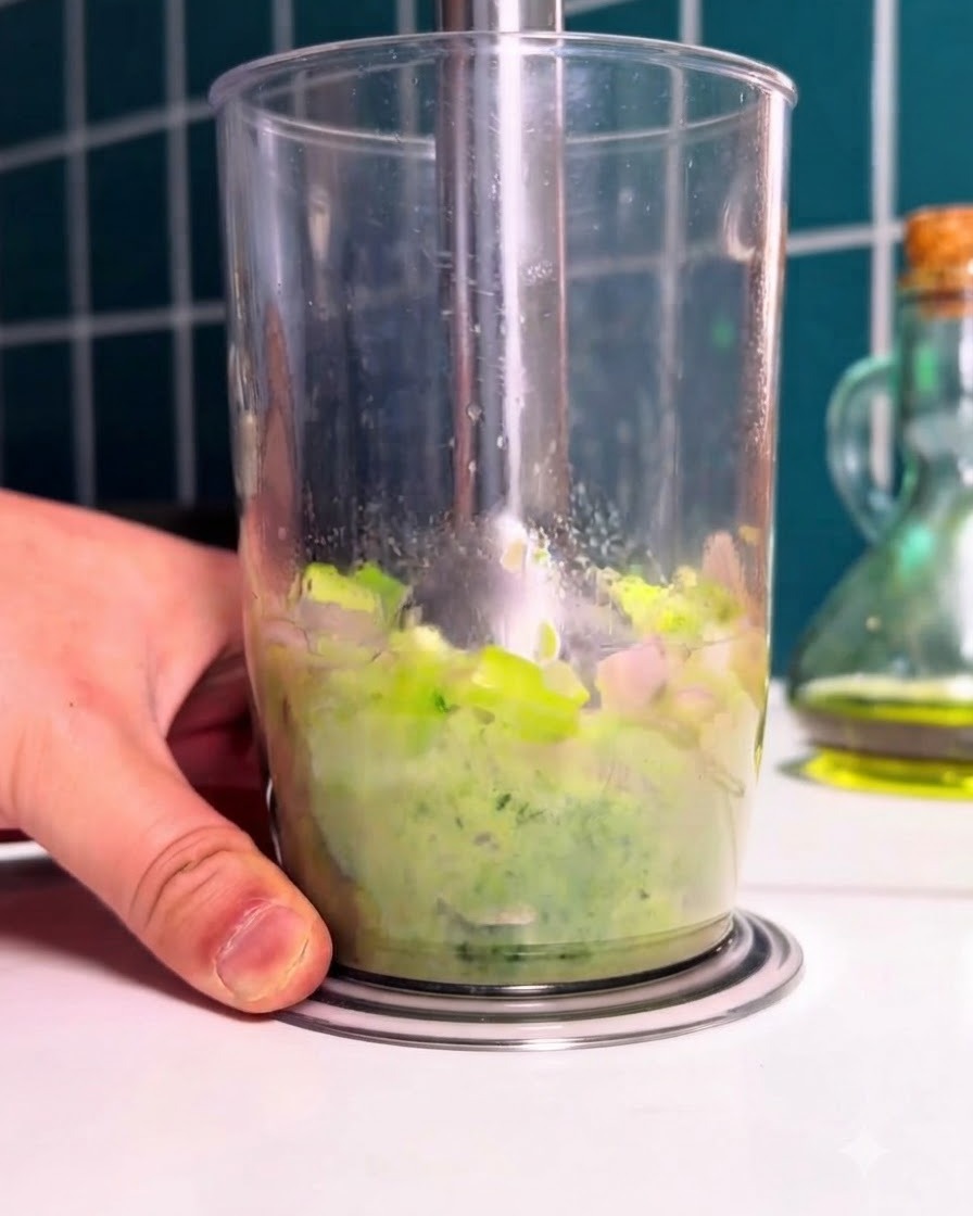 Blender ręczny pureeujący zblansowane zielone warzywa i szpinak wewnątrz wysokiego przezroczystego kubka.