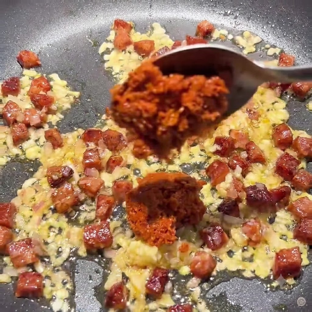 Łyżka dodająca gęstą pastę z suszonych pomidorów i przecier pomidorowy na patelnię z mieszaniną chorizo i cebuli.