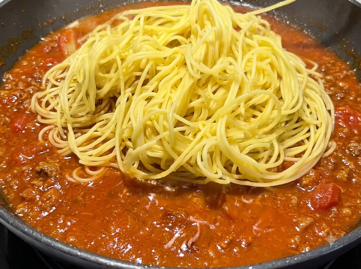 Kopiec ugotowanego żółtego spaghetti umieszczony na wierzchu duszącego się czerwonego sosu na patelni.