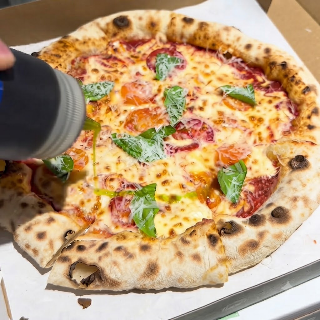 Butelka nalewająca strumień oliwy z oliwek z pierwszego tłoczenia na świeżo upieczoną pizzę polaną bazylią i serem w pudełku.