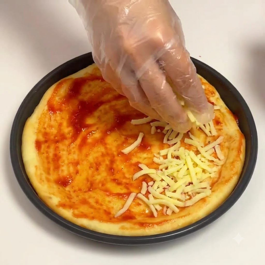 Ręka w rękawiczce posypująca startym serem mozzarella bazę z sosu pomidorowego w okrągłej formie do pizzy.