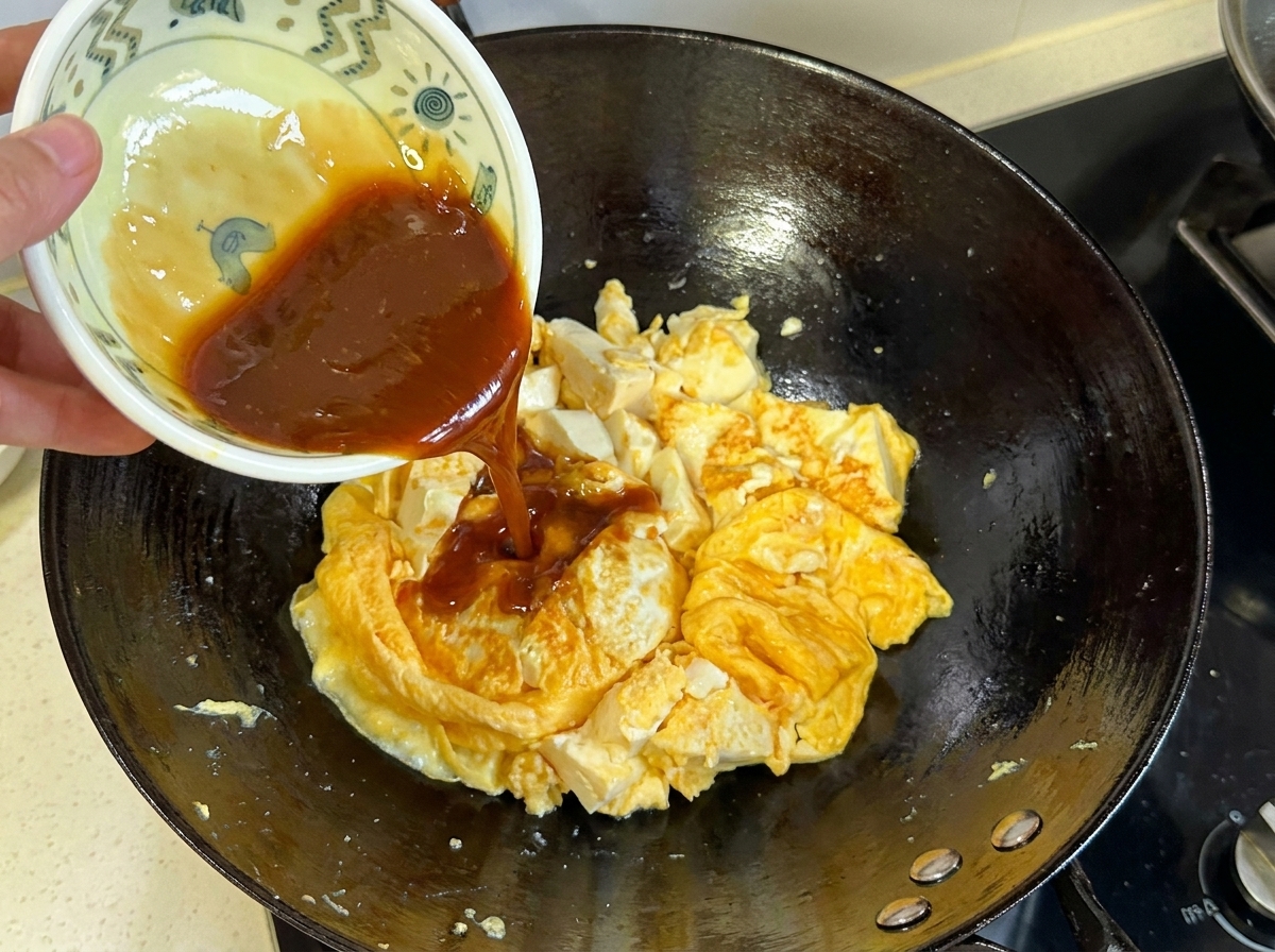 Ciemnobrązowy sos wlewany z małej miseczki na podsmażoną mieszankę jajka i tofu w woku.