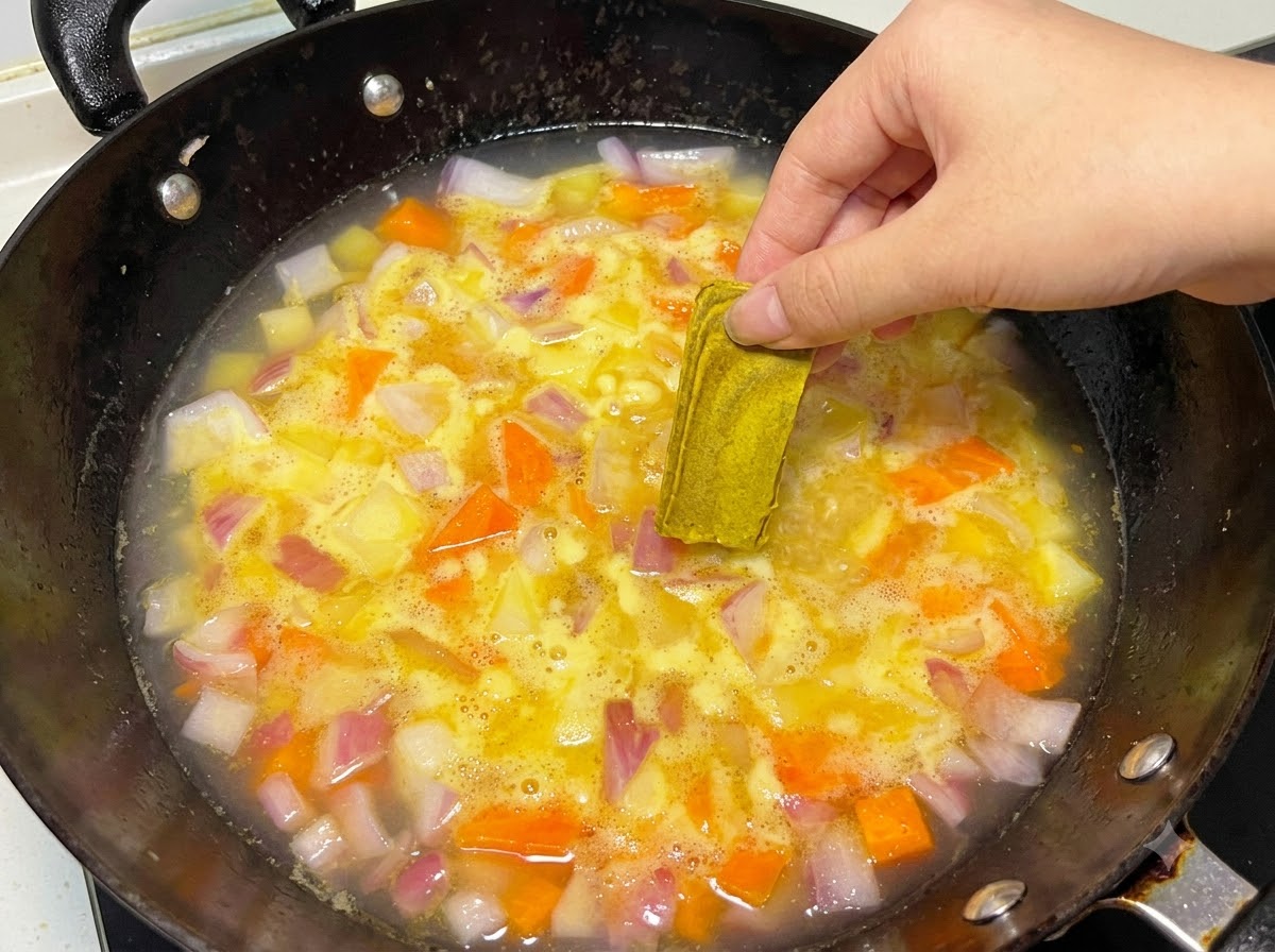 Ręka umieszczająca solidną kostkę curry w gotującym się garnku z wodą i warzywami.