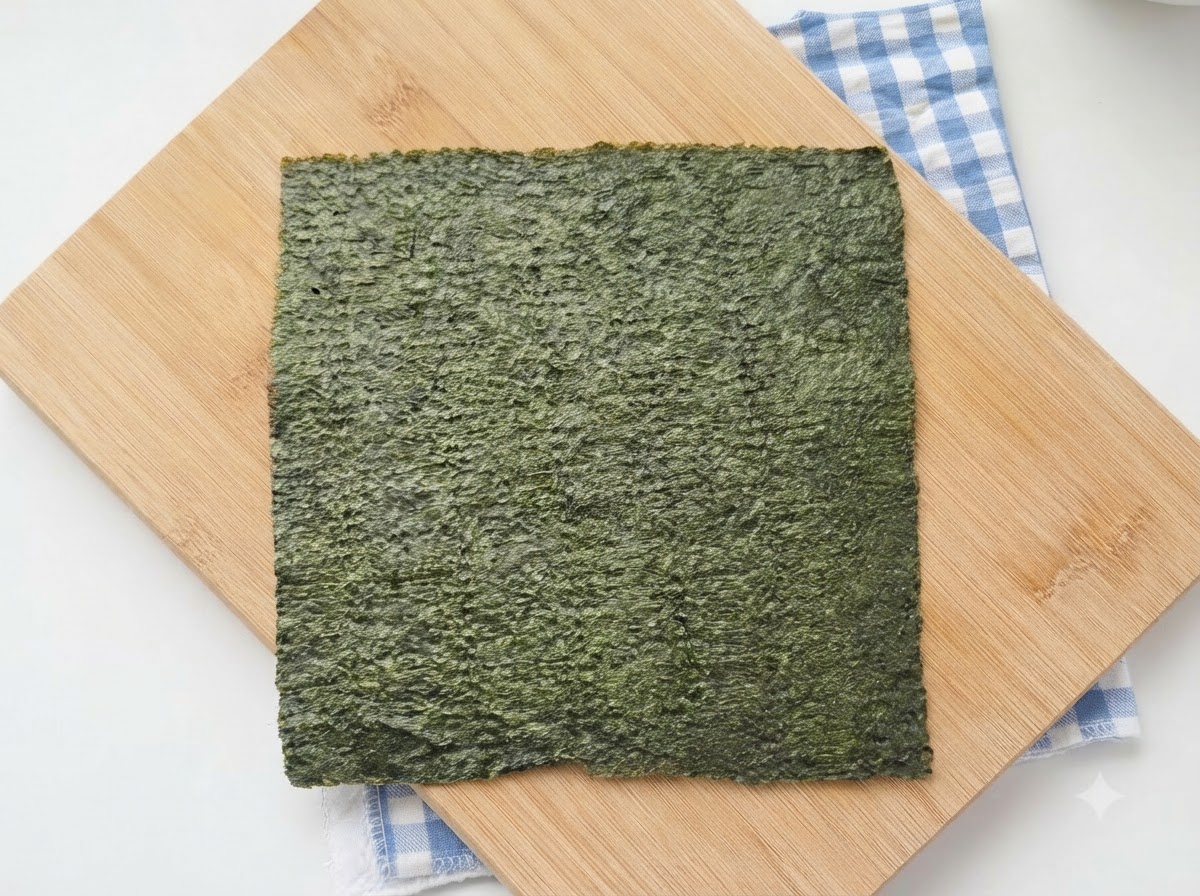 Pełny płat zielonych alg nori do sushi leżący na drewnianej desce do krojenia, pod którą znajduje się niebieska kraciasta ściereczka.