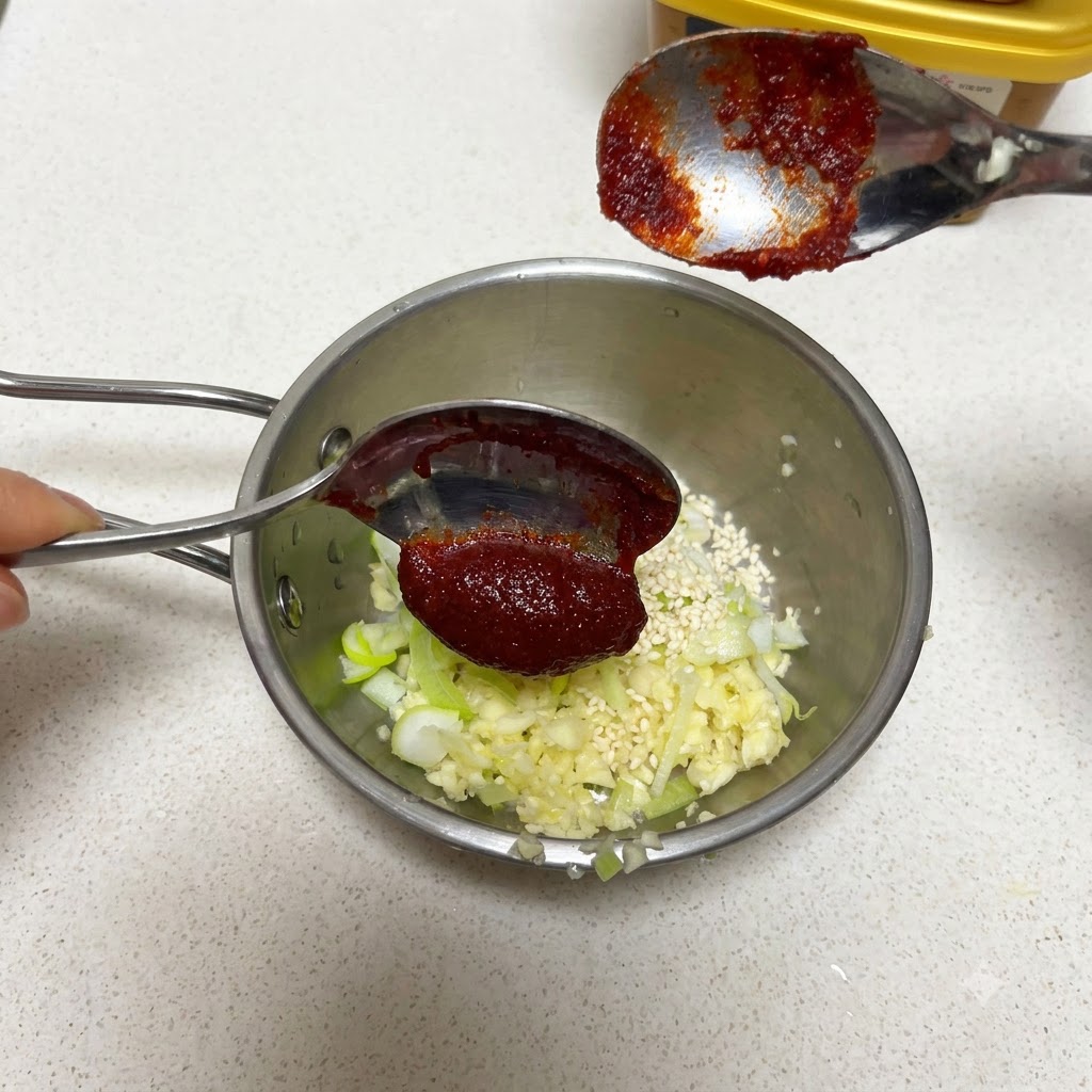 Łyżka koreańskiej pasty chili trzymana nad patelnią z czosnkiem i nasionami sezamu.