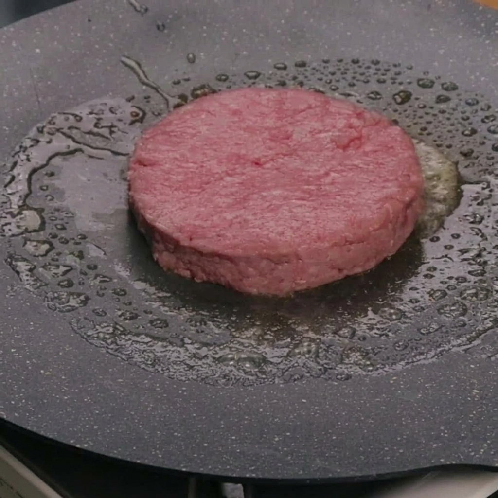 Surowy kotlet wołowy smażący się na maśle na rozgrzanej patelni.