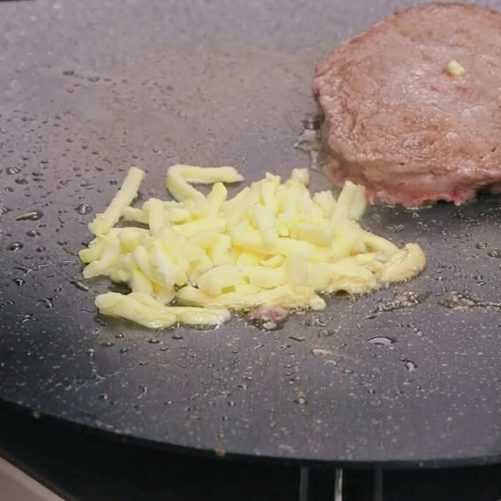 Starty ser mozzarella topiący się na wierzchu podsmażonego kotleta wołowego na gorącej patelni.
