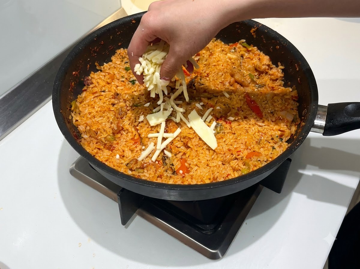 Ręka posypująca obfite ilości startego sera mozzarella na patelnię z czerwonym, pikantnym smażonym ryżem.