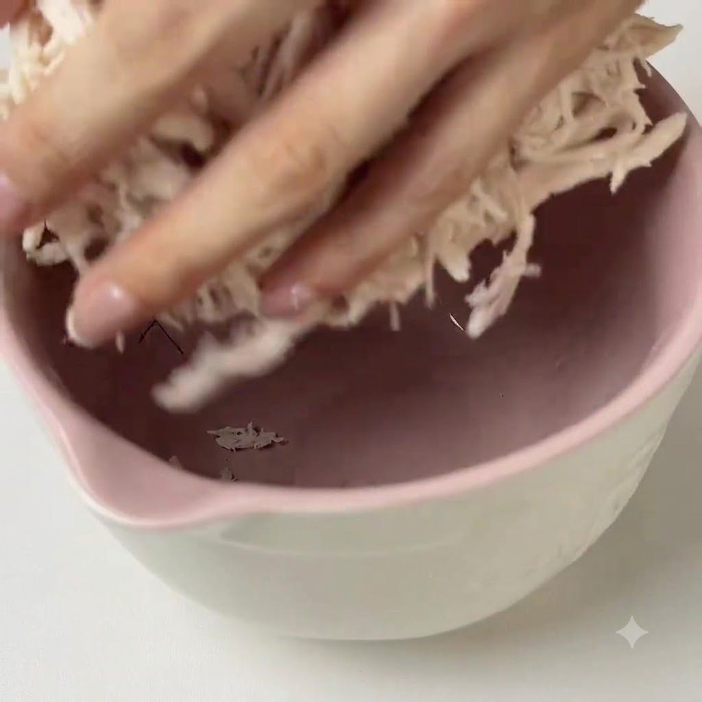 Ręce mieszające zielone pesto z poszarpanym kurczakiem w różowej misce.